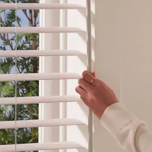 Close-up of hand opening or closing white window blinds, natural light entering, outdoor scenery visible through slats, home interior, window treatment, sunlight control, privacy, modern decor.