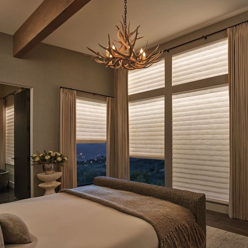 Elegant bedroom with large windows featuring white pleated shades and cream curtains, creating a warm and inviting atmosphere with soft lighting.