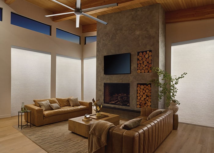 Spacious living room featuring a contemporary stone fireplace, high ceilings with wooden beams, large windows with blinds, and cozy leather sofas. Elegant, bright, with natural lighting.