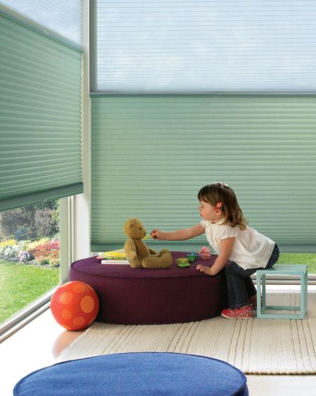 A young girl reaches out to a teddy bear while seated on a purple ottoman in a sunny playroom with large windows and green blinds.