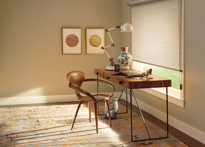 A stylish home office featuring a wooden desk, modern chair, decorative vase, adjustable desk lamp, and a large window with a beige roller shade, creating a bright and inviting workspace.