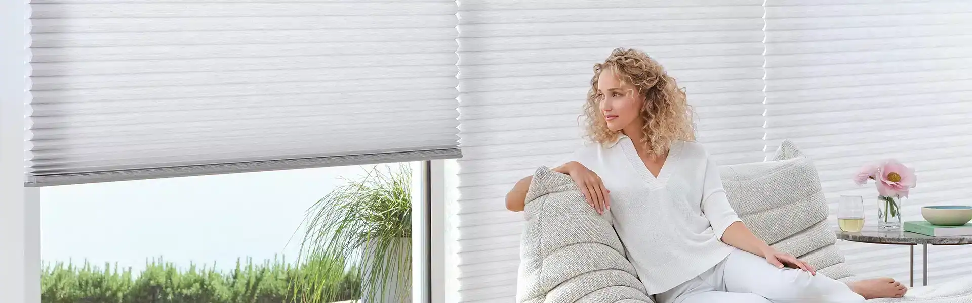 Bright living room with large window featuring white horizontal blinds, cozy sofa, and decorative plants, creating a welcoming and stylish space.