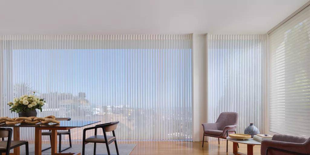Spacious living space with large windows featuring vertical blinds, minimalist furniture, cozy armchair, and decorative accents, perfect for stylish, natural light-filled interiors.