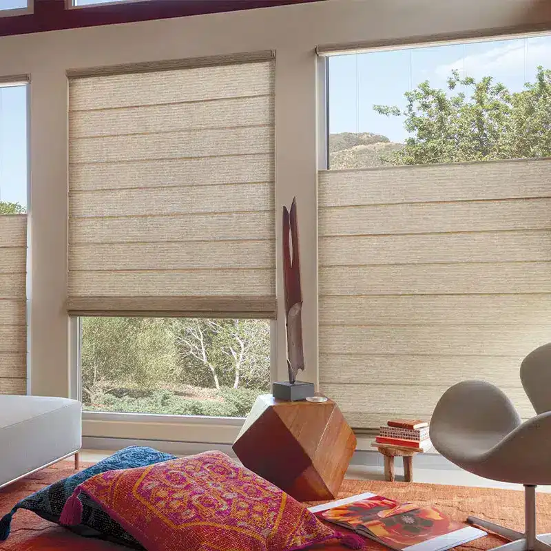 Beautiful image of beige window shades partially rolled down in a cozy, modern living room with natural light, greenery outside, and stylish decor including books, a chair, and artwork.