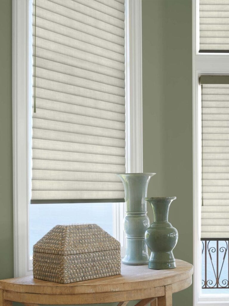 Window blinds in a cozy room with natural light, featuring a beige color and horizontal stripes. The scene includes stylish ceramic vases and a decorative box, creating a warm and inviting interior design setting.