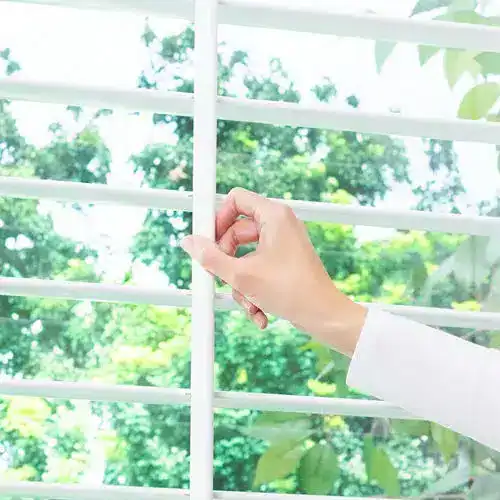 Close-up of a hand adjusting white window blinds with sunlight and greenery outside, emphasizing window treatment and natural light in home decor.