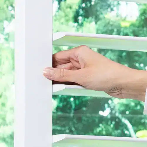 Close-up of a hand adjusting a white vertical louver vent cover on a window with a green outdoor background.