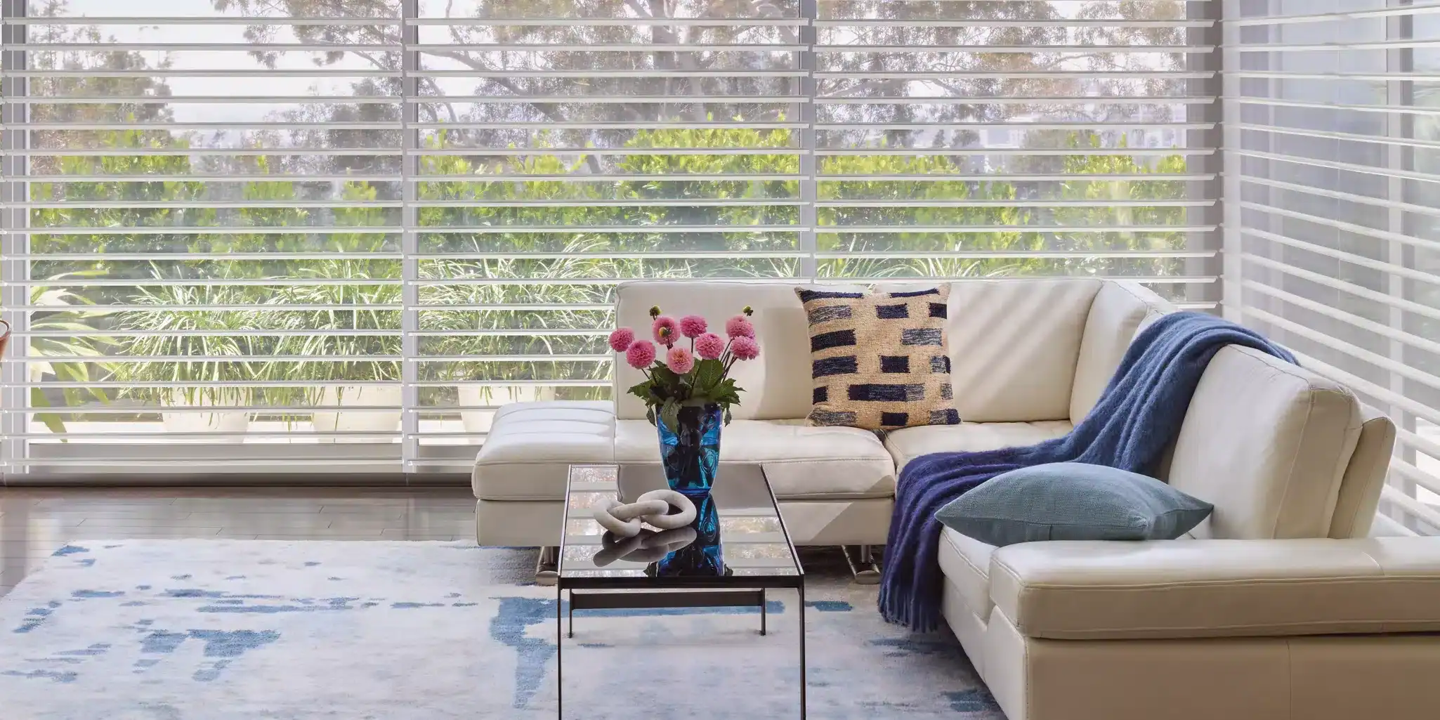 Vivid pink flowers in a blue glass vase on a modern glass coffee table in front of a neutral-colored sectional sofa with decorative pillows and a cozy throw blanket, set against large windows with white horizontal blinds.