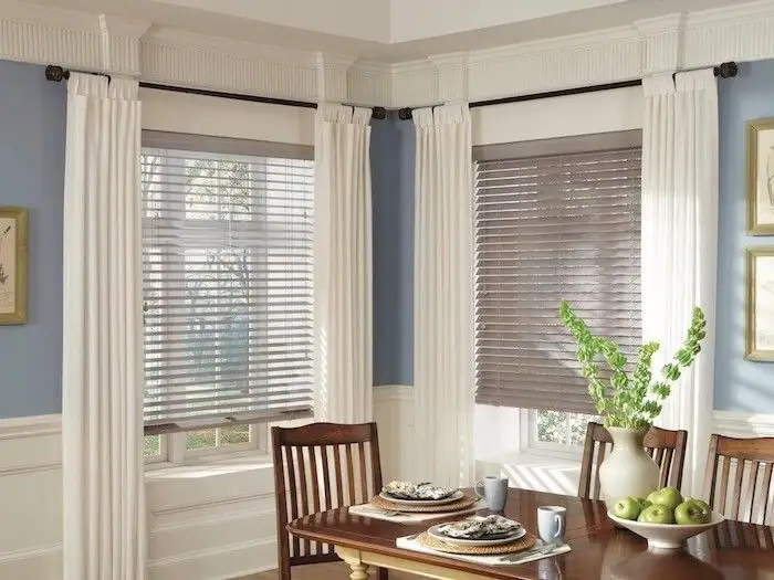 Living room with large windows, white curtains, and brown blinds, featuring a wooden dining table with chairs, a vase with green plants, and framed artwork on the walls.