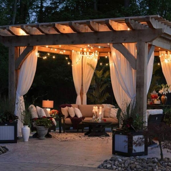 Cozy outdoor lounge under a wooden pergola with string lights, curtains, and comfortable seating for evening relaxation.