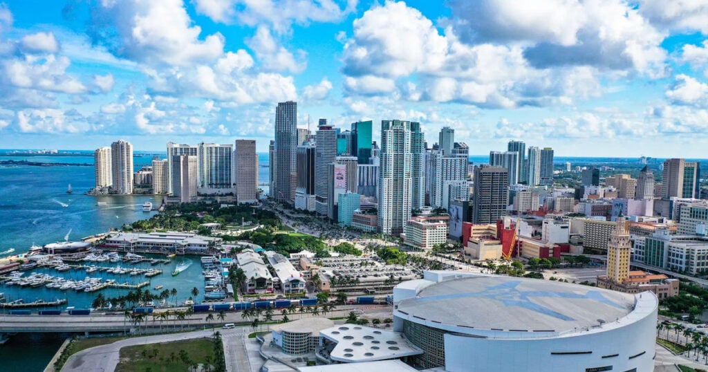 Aerial view of Miami-Dade County skyline with high-rise buildings, waterfront, and cruise port under a partly cloudy sky.