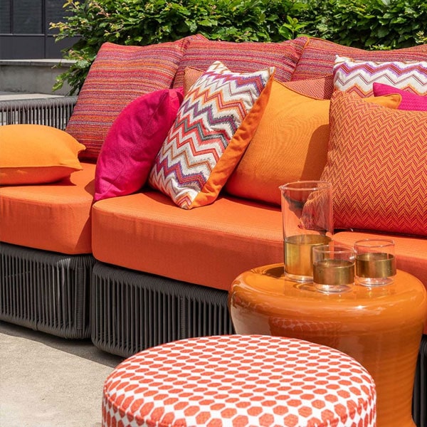 Vibrant outdoor sofa with orange, pink, and patterned cushions, complemented by a matching ottoman and decorative glassware, set against lush greenery.